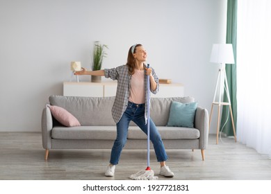 House cleaning with fun. Happy housewife singing her favorite song during cleanup, using mop as microphone, enjoying domestic work. Positive young maid tidying her home with pleasure