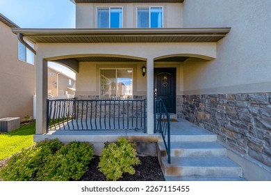 House entrance at the side of a wall with stucco and stone veneer. House exterior with railings on the porch at front of the window beside the black front door with hanging ornaments. - Shutterstock ID 2267115455