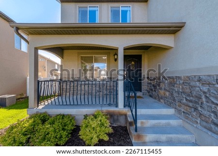 House entrance at the side of a wall with stucco and stone veneer. House exterior with railings on the porch at front of the window beside the black front door with hanging ornaments.