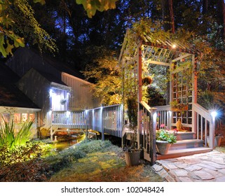 House Exterior With Porch Walkway Over A Pond And Lighting In The Woods