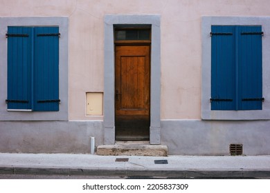 House Facade With A Dark Brown Wooden Door And Two Closed Blue Wooden Windows. Frontal View With Sidewalk. No People. Traditional House In An Old Town Of France