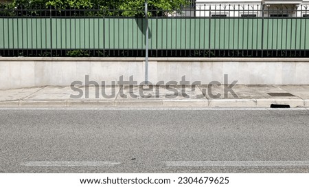 House fence consisting of a low concrete wall with metal railing and a green plastic panel. Cement sidewalk and asphalt street in front. Background for copy space.