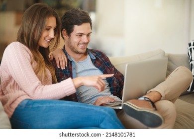 House Hunting From The Comfort Of Their Couch. Shot Of A Happy Young Couple Using A Laptop Together While Relaxing On The Sofa At Home.