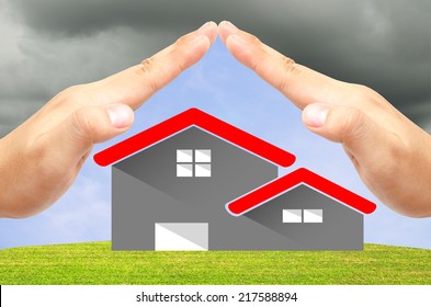 House Icon On The Green Field And Blue Sky Background Under 2 Open Hands Attach Together Like A Roof For Protection It From The Storm.