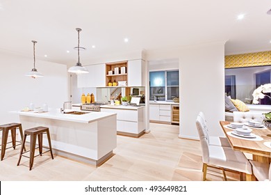 House Interior With White Walls, Focusing The Kitchen And Dining Area With Wooden Table Set Up Beside A Small Bed And Pillow. There Are Hanging Lights In The Kitchen With A Silver Stove