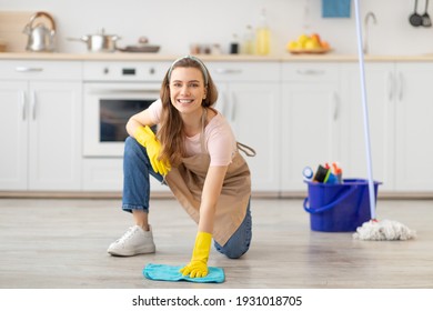 House keeping concept. Happy young woman wiping floor in kitchen, wearing rubber gloves, using cleaning supplies. Lovely housewife looking at camera and smiling, copy space