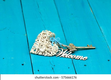 House Key Attached To Natural Wicker Heart With Ribbon And Welcome On Antique Rustic Teal Blue Wood Background; New Home, Valentines Day, Love And Welcome Home  Background With Painted Copy Space