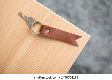 A House Key With A Leather Brown Key Tag Attached To It. Antique Brass Hardware Such As The Rivet And Key Ring. Laying Down On A Natural Wooden Table With Grey Carpet In The Background