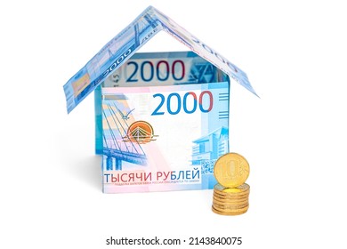 A house made of ruble notes and a stack of coins isolated on a white background. Text translation: 2000 thousand rubles. Forgery of Bank of Russia tickets is prosecuted by law