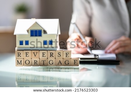 House Model Over Reverse Mortgage Blocks In Front Of Businesswoman Writing On Paper Over Desk