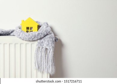 House model wrapped in scarf on radiator indoors, space for text. Winter heating efficiency