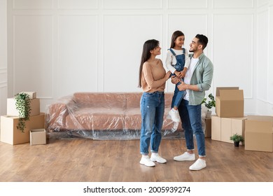 House Move, Repair Concept. Full Body Length Of Cheerful Young Family Of Three People Standing In New Empty Apartment, Looking At Each Other, Free Copy Space. Parents Holding Daughter On Hands