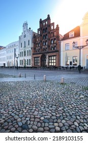 House The Old Swede In Wismar, Germany, With Other Historic Buildings. Empty Square Early Morning, With Flare. Sunny Day With Blue Sky.