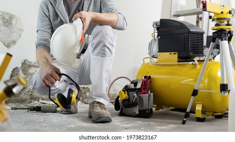 house renovation concept, construction worker holds in hands helmet and safety headphones, air compressor and construction tools in the background