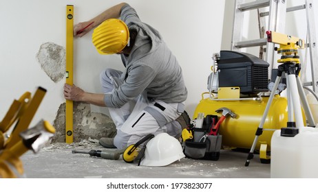 house renovation concept, man worker looks at the spirit level checks the wall, helmet and safety headphones, air compressor and construction tools in background