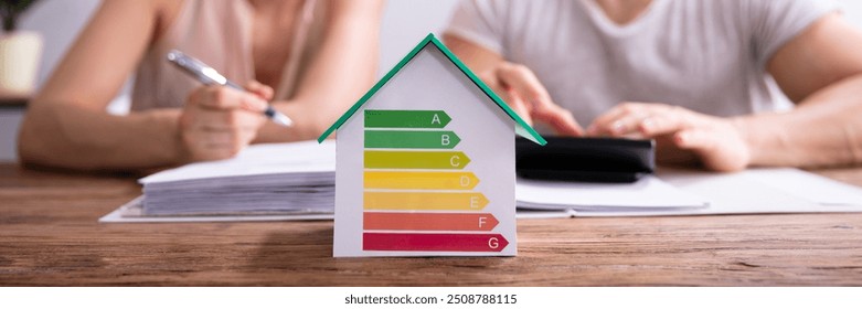 House Showing Energy Efficiency Rate In Front Of Happy Couple Using Calculator Over Wooden Desk - Powered by Shutterstock