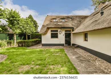 a house with thatched roof and stone pathway leading to the front door, surrounded by lush green trees on a blue sky day