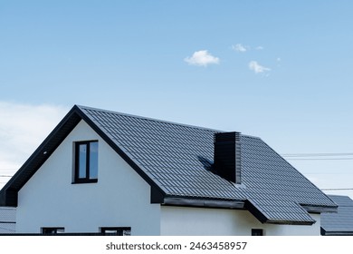 A house with white siding and a black roof is set against a blue sky with fluffy white clouds. The engineering of the sloped roof and facade windows adds character to the building - Powered by Shutterstock