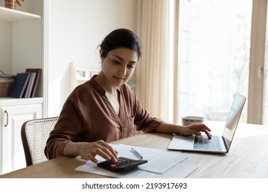 Household budget. Young Indian female sit at table calculate utility payments before make money transfer in online bank app on laptop. Focused millennial business woman pay taxes plan personal
