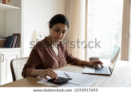 Household budget. Young Indian female sit at table calculate utility payments before make money transfer in online bank app on laptop. Focused millennial business woman pay taxes plan personal