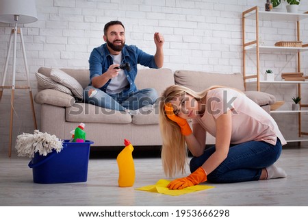 Household chores, fatigue and chauvinism. Sad young woman housewife in rubber gloves washes floor with cleaning supplies, horny guy plays in online game with joystick on sofa at home, free space