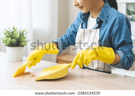 Household clean up, housekeeper asian young woman wearing protection rubber yellow gloves, using broom, dustpan sweeping remove spilled crumb food broken on dirty table at home, equipment for cleaning