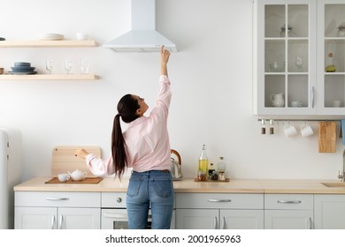 Household Concept. Back Rear View Of Casual Lady Choosing And Selecting Mode On Range Exhaust Hood, Pushing Button On Mechanical Fan Above The Stove. Female Standing Cooking In Modern Kitchen At Home