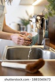 Household And Eco Friendly Supplies Concept. Vertical View Of Woman Standing On Kitchen, Washing Bowl With Zero Waste Sponge While On Front Blurred Background Lying Wooden Coconut Brush