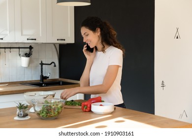 Household Multitask. Active Young Housewife Preparing Food At Kitchen Communicating By Cell Holding Device At Ear. Happy Millennial Woman Engaged In Cooking Process Talk Chat On Phone In Good Mood