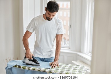 Housekeeping and housework concept. Hard working handsome husband irons clothes on ironing board, does household chores, helps wife, stands indoor near window, dressed in white casual t shirt - Shutterstock ID 1169708104