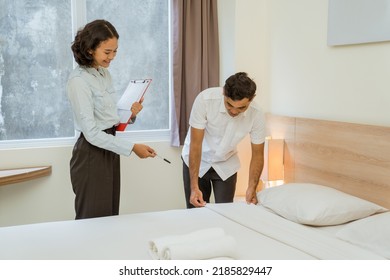 Housekeeping Manager Carrying Clipboard Notes While Supervising Maid Cleaning Bed In Hotel Room