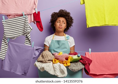 Housekeeping And Washing Concept. Dissatisfied Sad Young Woman Has Afro Hairstyle, Hangs Clothes On Clothes Lines With Clips, Does Laundry At Home, Wears Casual Apron, Looks Aside, Stands Indoor