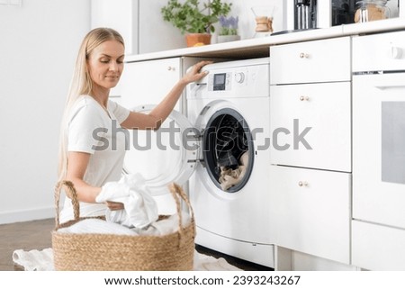 housekeeping woman doing laundry at home. smiling female takes clothes from wicker basket and put textile in automatic washing machine with open door. built in appliance on kitchen with light interior
