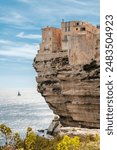 Houses perched on limestone cliffs undercut by the sea and weather in the citadel of Bonifacio in Corsica with a sailing yacht in the distance