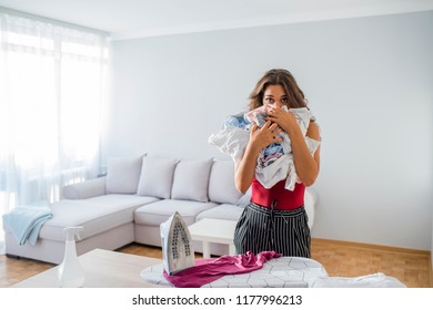 Housewife Bringing A Huge Pile Of Laundry On The Ironing Board, Boring Household Chores Concept. Disappointed Unhappy Housewife. Sad Woman Ironing Clothing At Home