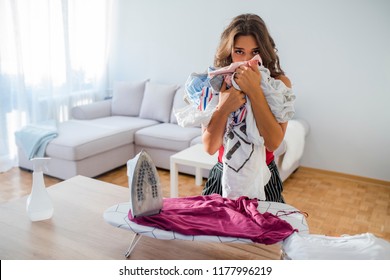 Housewife Bringing A Huge Pile Of Laundry On The Ironing Board, Boring Household Chores Concept. Disappointed Unhappy Housewife. Sad Woman Ironing Clothing At Home