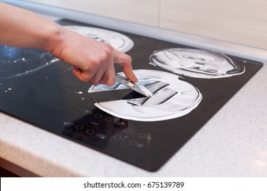  Housewife cleaning and polish electric cooker. Black shiny surface of kitchen top, hands with foam, glass scraper, bottle of cleaning agent
