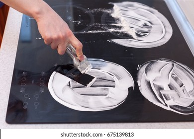 Housewife cleaning and polish electric cooker. Black shiny surface of kitchen top, hands with foam, glass scraper, bottle of cleaning agent