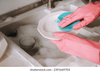 housewife girl in pink gloves washes dishes by hand in the sink with detergent. The girl cleans the house and washes dishes in gloves at home - Powered by Shutterstock