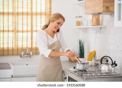 Housewife on a call. Blonde woman stirring something in a saucepan and talking on the phone