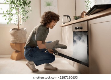 Housewife opening the oven door and taking out burnt food during her cooking in the kitchen