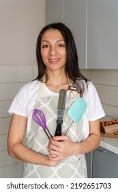 Housewife Is Standing In The Kitchen With A Large Pot And A Whisk. Confectioner Woman In Apron Preparing To Cook. Woman Wearing Apron And Mixing Batter. Cooking Concept With Professional