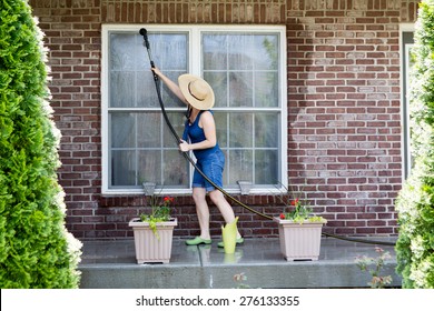 Housewife Standing On A Patio Washing The Windows Of Her House With A Hose Attachment As She Spring-cleans The Exterior At The Start Of The New Spring Season