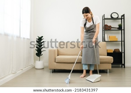 Housewife using broom and dustpan wearing an apron to clean the living room at apartment. Young woman is happy to clean home. Maid cleaning service.
