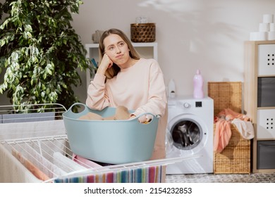 Housewife Woman Tired Of Household Duties. Girl In Sweatpants Stands In Laundry Room Next To Clothes Dryer, Props Up Chin Thoughtful Sad Looks Out Window At Sunny Weather, Worried, Disgusted.