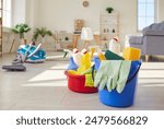 Housework equipment. Blue plastic bucket and red basin filled with yellow white detergent bottles, rubber gloves, sponges and other cleaning tools, on floor at home, with vacuum cleaner in background