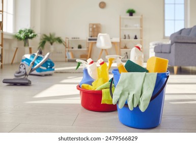 Housework equipment. Blue plastic bucket and red basin filled with yellow white detergent bottles, rubber gloves, sponges and other cleaning tools, on floor at home, with vacuum cleaner in background - Powered by Shutterstock