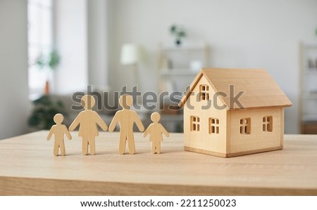 Housing for families. Close up of wooden figurines of family with children and model of small one-story house. Miniature figurines made of light wood stand on table in bright room. Blurred background.