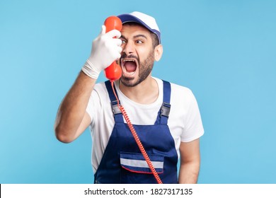 Housing maintenance call centre. Crazy handyman in overalls, safety gloves, shouting into retro phone handset, irritated by customer call. Plumber and repair services. indoor studio shot, isolated