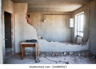 Housing redevelopment. Small table with perforator, sledgehammer placed on floor at partially destroyed inner wall. On backdrop empty walls, window, gas boiler, meter and pipes connecting them.